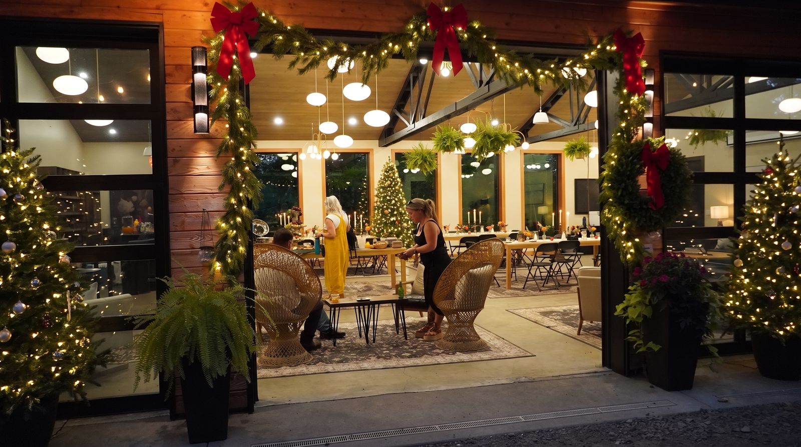 Dusk at the hall with its glass garage door rolled open, framed in lit pine garland and red bows, a lit tree on each side and guests inside